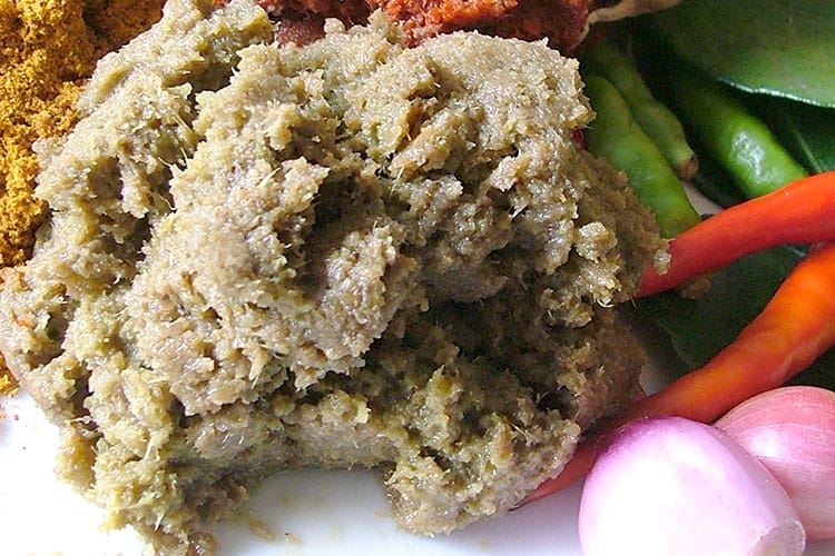 A portion of freshly made green chili paste next to green chilis and shallots, with two red chilis on the side and curry powder and red chili paste in the background.