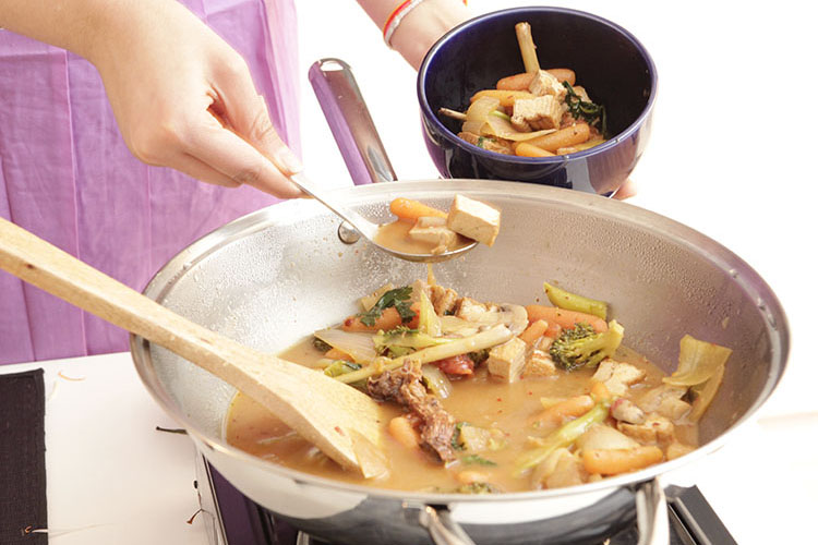 Student wearing a lavendar apron, spooning completed Tom Yam soup recipe made with fresh chili paste from stainless steel wok into a dark blue ceramic bowl using a stainless steel spoon. A large wooden spoon rests in the wok with large piecs of broccoli, onion, tofu and soup broth.