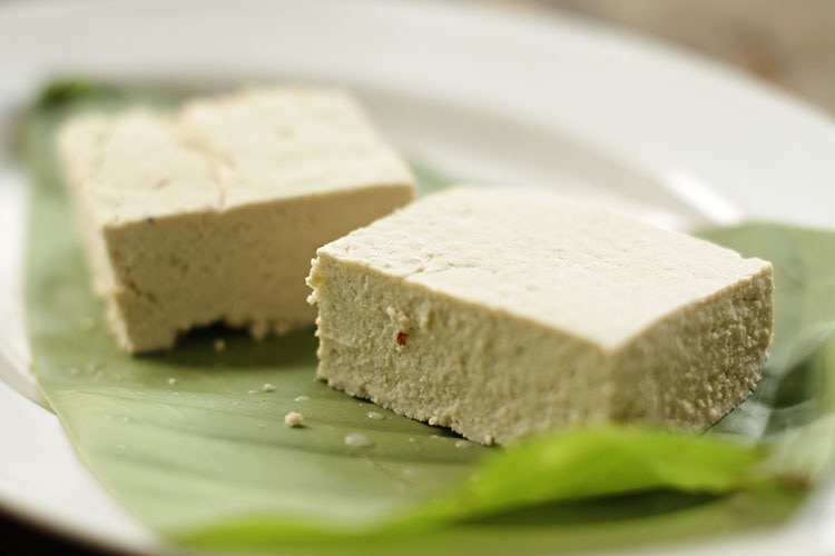 Two freshly made cut blocks of tofu placed on a banana leaf on a white plate.