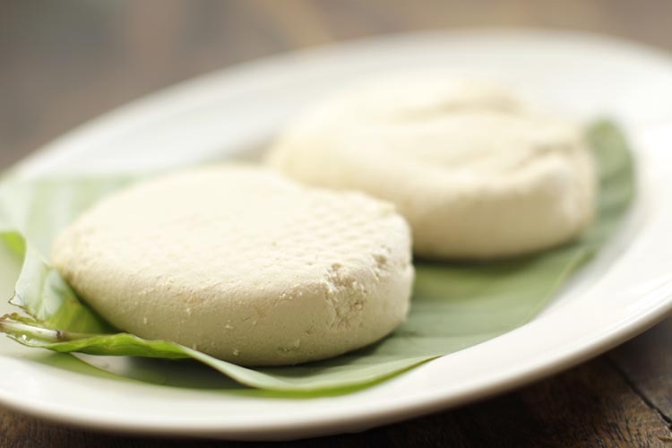 Final rounded blocks of pressed freshly made tofu sitting on a banana leaf on top of a white plate.