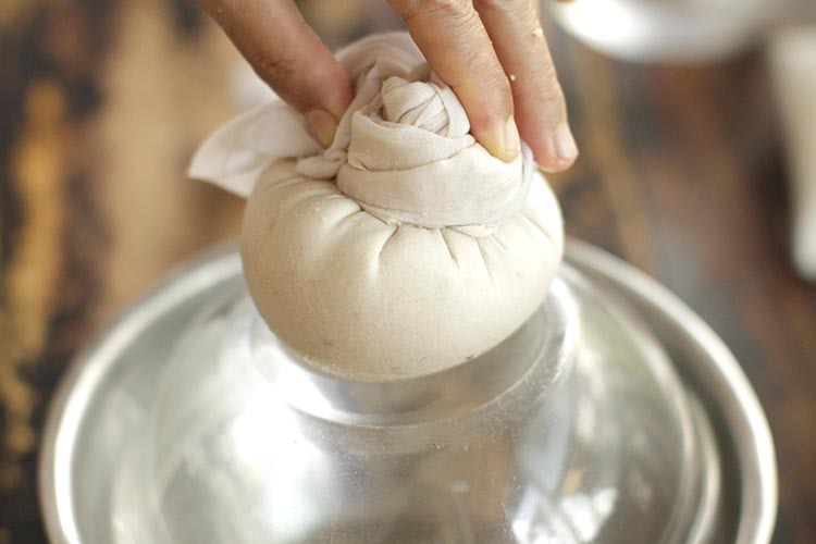Fingers holding down tofu in cheesecloth on an upside down stainless steel bowl, squeezing remaining soy milk from freshly made tofu.