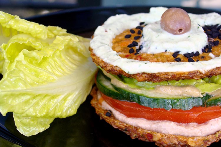 A vegan Raw burger with sauce with cream sauce, lettuce, cucumber and tomato slices and lettuce on the side.