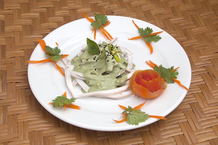 Vegan Raw pad thai topped with a basil cream sauce on a white plate with carrot, parsley and a tomato fruit carving garnish.