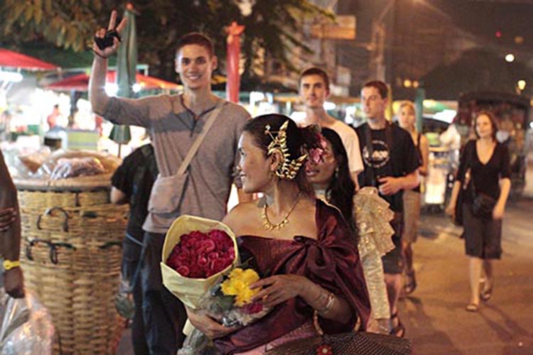 Group of students, three women and three men, at flower market in Bangkok. The man in the center and woman on the right holding a bouquet of pink, yellow and white flowers.