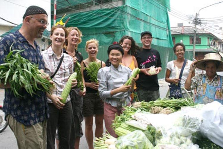 Students standing with the founder of May Kaidee holding produce next to a Bangkok vegetable vendor.