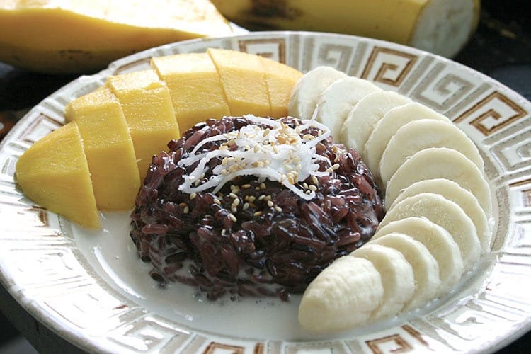 Black sticky rice and mango dessert. Slices of mango and banana carefully arranged around a mound of black sticky rice topped with shredded coconut pulp and sesame seeds.