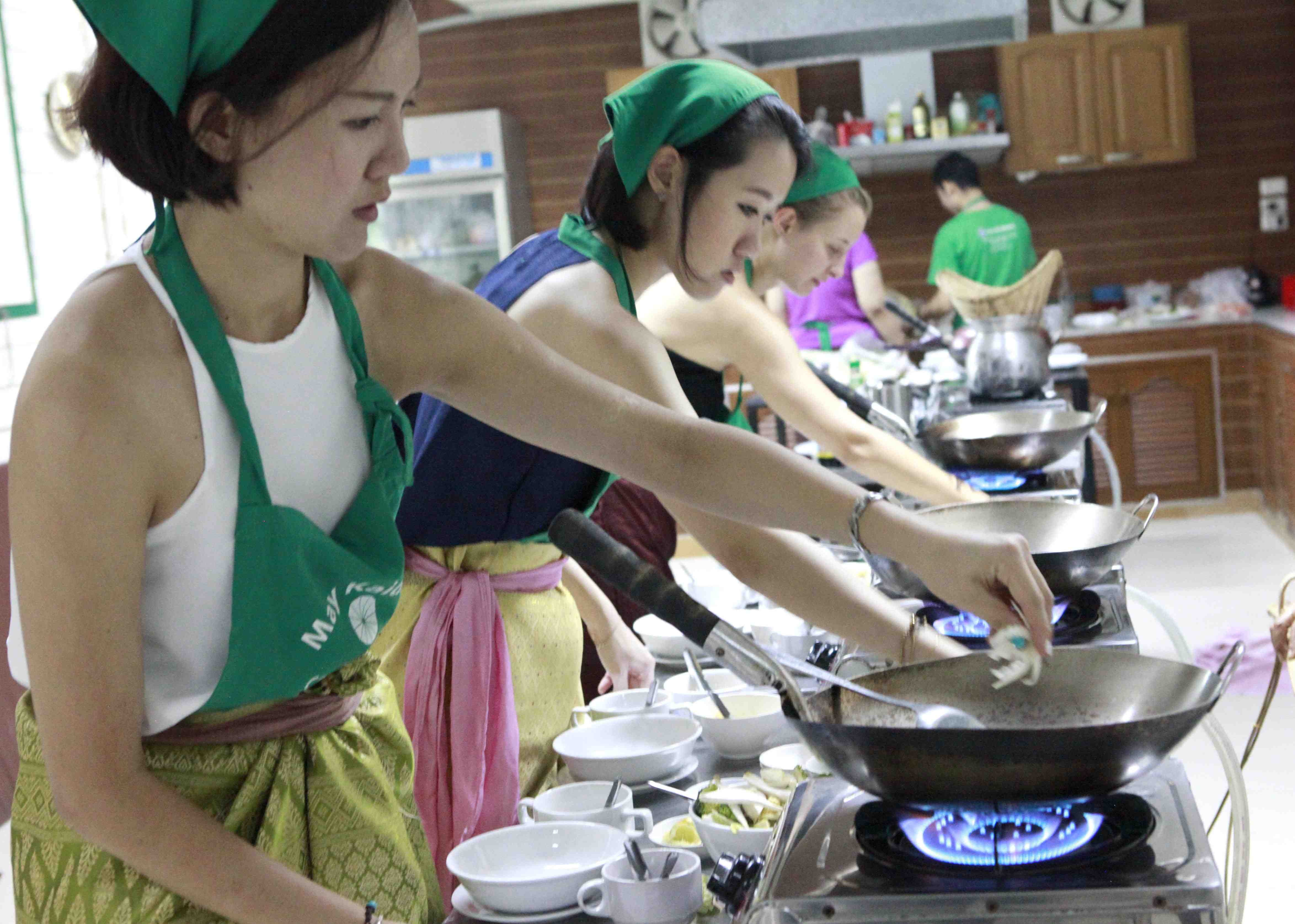 Students with green aprons and headscarves cooking Thai food on gas woks.