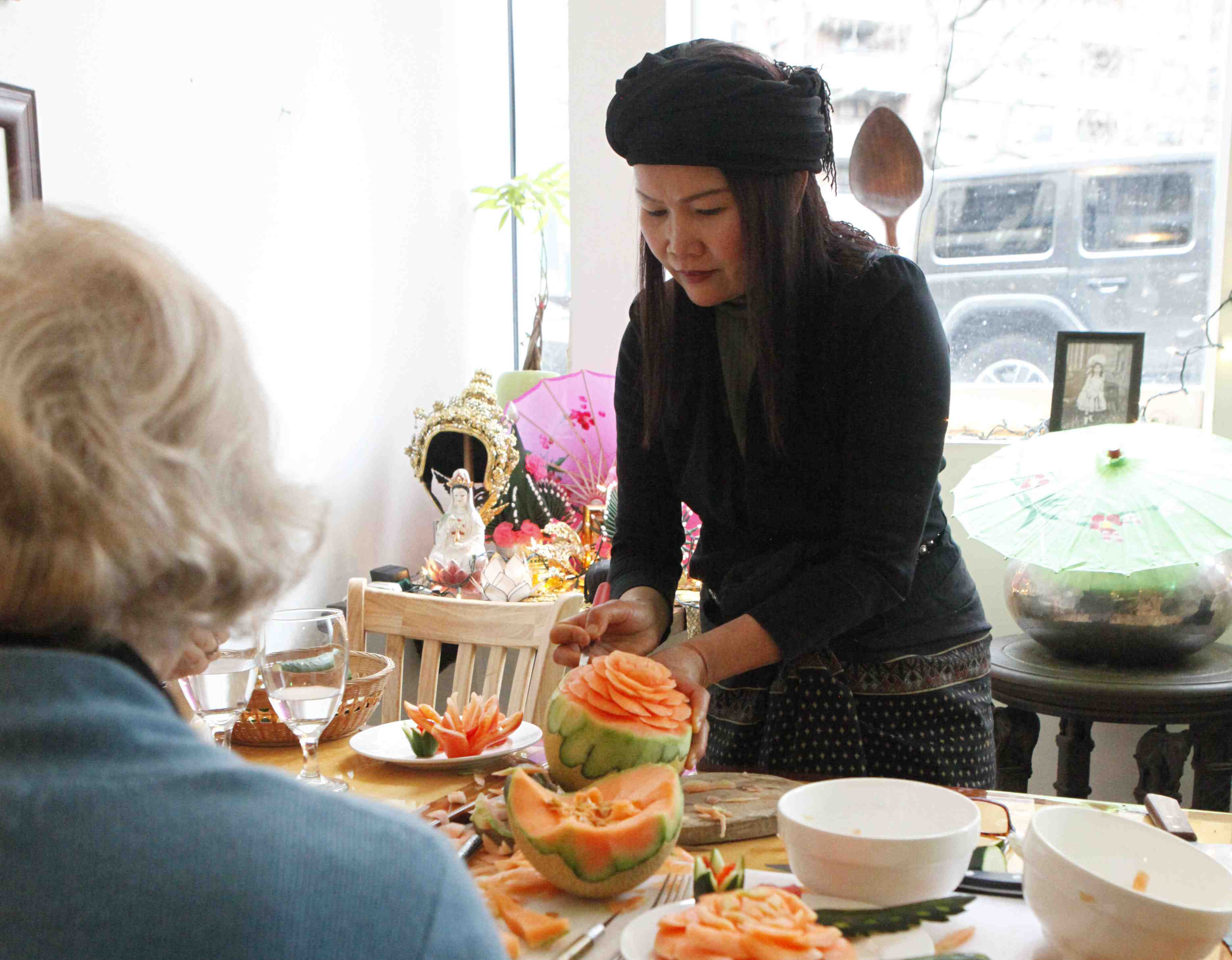 A fruit carving student patiently watching an instructor carve a cantaloupe into a flower design. Other cantaloupe carvings sitting on the table surrounded by additional decorations.