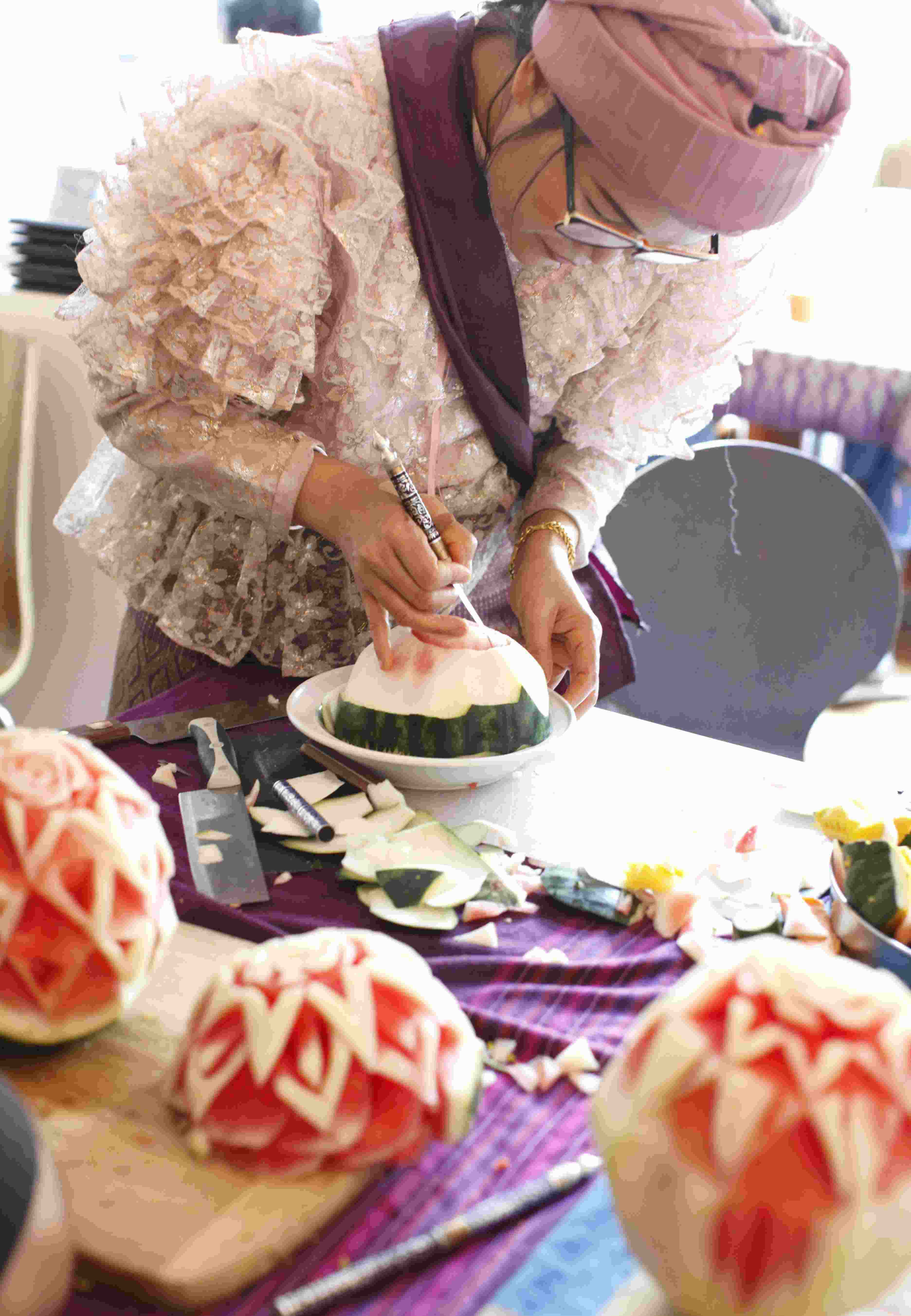 A fruit carving instructor demonstrating how to carve a watermelon into a large floral design on a table with a Thai tablecloth, three other carved watermelons, carving knives and assorted pieces of fruit carving pieices.