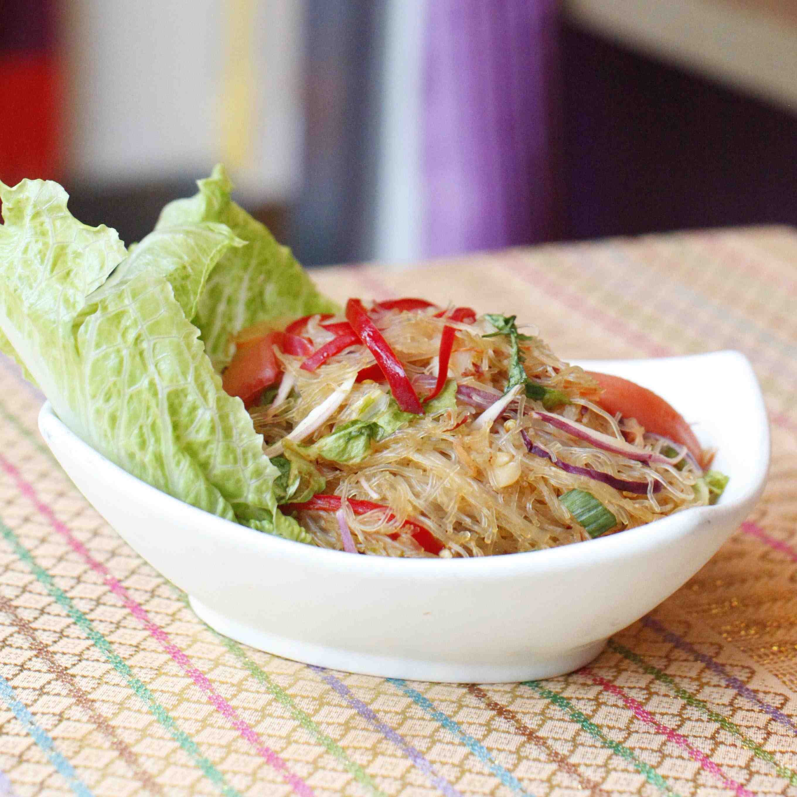 Glass Noodle Salad, made of mung bean noodles soaked in water then steamed and cooled, slices of red bell pepper, toasted rice powder and chopped mint leaves served on large lettuce leaves in a white ceramic bowl.
